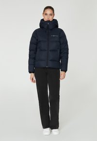 Marineblaue Daunenjacke mit hohem Kragen und Kapuze, ausgestattet mit einem Frontreißverschluss, gestepptem Material und einem Logo auf der linken Brust. Kombiniert mit schwarzen Jeans.