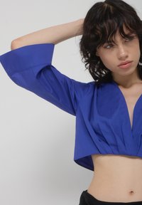 Blusa blu corta con maniche a sbuffo, caratterizzata da un profondo scollo a V e dettagli arricciati nella parte frontale. Tessuto morbido e leggero.