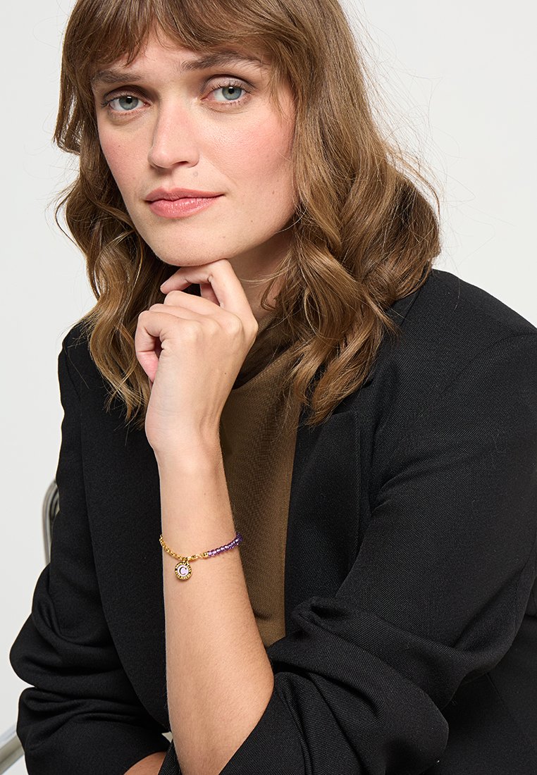 Gold- und lila Armband mit Perlen und einem runden Anhänger; das Model stützt ihr Kinn auf die Hand, trägt ein schwarzes Blazer und ein braunes Oberteil.