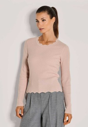 Vrouw met donker haar in een paardenstaart, gekleed in een lichtroze geribbelde lange mouwtop met gegolfde randen en grijze high-waisted broek.
