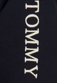 Sudadera de color azul marino con texto bordado en crema en vertical "TOMMY." Textura suave, sin patrones visibles ni detalles adicionales.