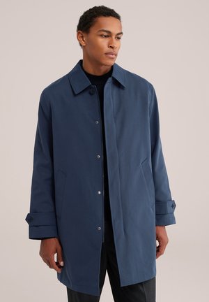 Jeune homme portant un manteau bleu foncé boutonné avec col et poches à rabat, debout devant un fond clair uni.