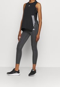 Černý bezrukávový sportovní top s vrstveným designem a bílými pruhy, spárovaný se šedými legínami a černými sportovními botami s bílými akcenty.