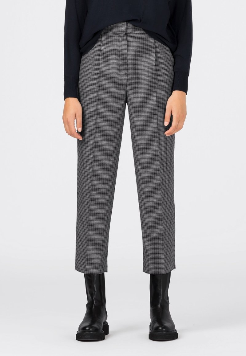 Personne portant un pantalon droit à carreaux gris, un pull noir et des bottines en cuir noires, debout sur un fond clair uni.