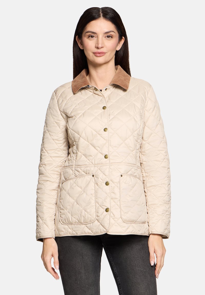 Femme portant une veste matelassée beige avec col marron, boutons-pression, et deux grandes poches avant, debout devant un fond blanc.