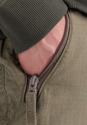 Hand teilweise in einer teilweise aufgezippte olivgrünen Jackentasche, getragen wird ein grünes langärmeliges, geripptes Oberteil.