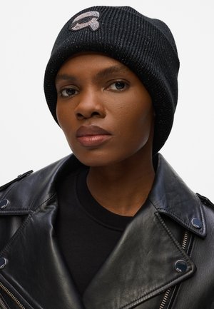 Black knitted beanie with ribbed texture and a decorative embroidered logo. Worn with a black leather jacket and crew-neck top.