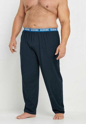 Hombre sin camiseta que lleva pantalones de pijama azul marino oscuro con cintura azul etiquetada "BADRHINO,", de pie descalzo contra un fondo liso.