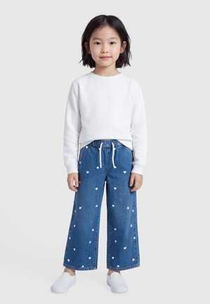 Joven chica con cabello negro que lleva una camiseta blanca de manga larga, vaqueros azules de pierna ancha con corazones blancos y zapatos blancos sin cordones, de pie sobre un fondo gris.