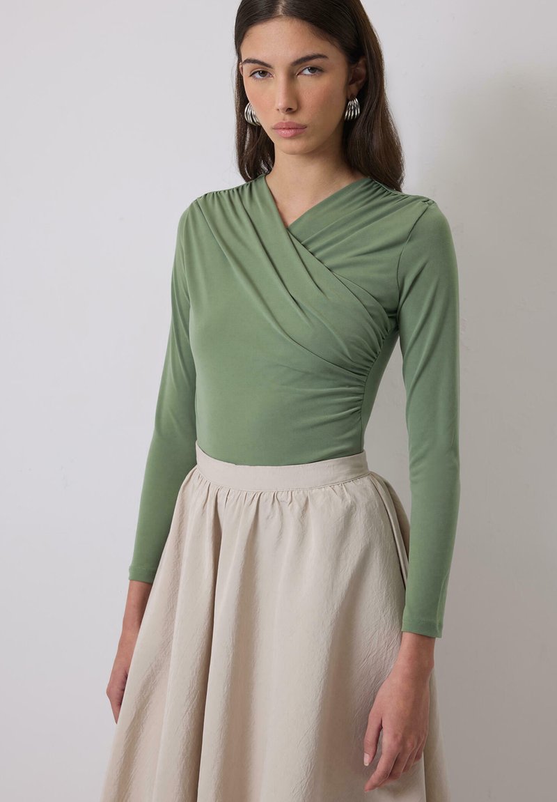 Green long-sleeve top with a wrap design and gathered detailing, paired with a beige knee-length skirt featuring a textured fabric.