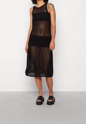 Femme portant une robe midi noire sans manches en maille transparente par-dessus des sous-vêtements noirs et des sandales à plateforme à bout ouvert noires, debout sur un sol gris avec un mur blanc en arrière-plan.