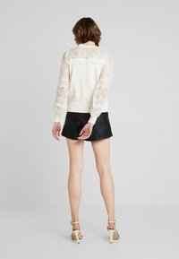 Pull à dos en dentelle avec corps crème et manches bouffantes, associé à une mini-jupe en cuir noire et des talons hauts beiges.