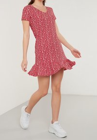 Rotes Kleid mit Blumenmuster, kurzen Ärmeln, ausgestelltem Saum und tailliertem Oberteil. Kombiniert mit weißen Sneakers. Glatter Stoff mit kleinen weißen Blumenmustern.