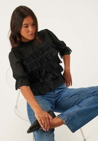 Blusa nera con dettagli a ruffle, maniche a palloncino e scollatura alta, abbinata a jeans blu a gamba dritta e ballerine nere. Design semplice.