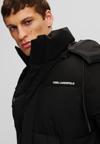 Čierna prešívaná bunda s vysokým golierom, podrobnosťami na zips a značkou Karl Lagerfeld. Obsahuje štruktúrovanú tkaninu a precízny dizajn.