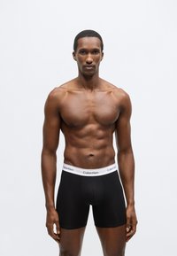 Boxeri negri din bumbac, cu talie elastică albă inscripționată cu "Calvin Klein", modelul stă cu o expresie neutră.