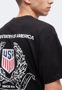 Άνδρας που φοράει μαύρο μπλουζάκι με το έμβλημα ασπίδας των ΗΠΑ, στεφάνι δάφνης και το κείμενο "United States of America" στην πλάτη, κοιτάζοντας πλάγια.