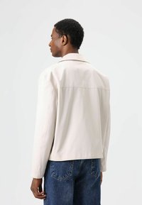 Lichtbeige jas met een gladde textuur, voorzien van een kraag en lange mouwen. Getoond vanaf de achterkant, gedragen over blauwe denim.