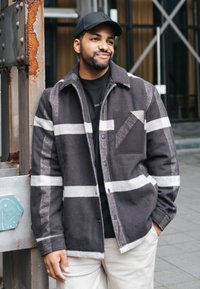 Muž opřený o zrezivělý kovový sloup, oblečený v černé kšiltovce, černobílé kostkované bundě, černém tričku a světlých kalhotách venku.