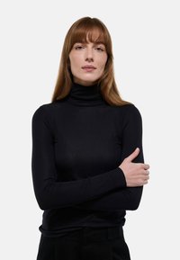 Haut à col roulé noir en tissu lisse avec des manches longues, à la coupe ajustée et à l'encolure discrète, porté avec les bras croisés.