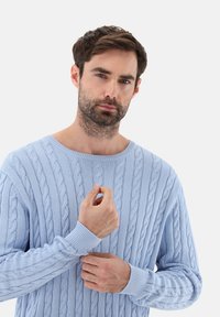 Hellblauer, strukturierten Muster versehener Strickpullover mit weitem Ausschnitt und gerippten Bündchen, vor einem schlichten weißen Hintergrund präsentiert.