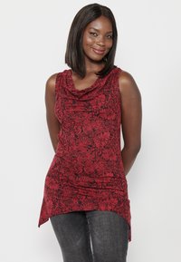 Haut sans manches rouge avec un motif floral, décolleté drapé et un ourlet arrière plus long, associé à un jean foncé. Tissu lisse avec un léger éclat.