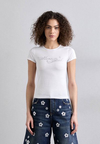 T-shirt bianco aderente con testo logo argento e maniche corte. Abbinato a jeans in denim blu decorati con applicazioni floreali.