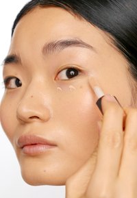 Gräddconcealer appliceras under ögat på slät, ljus hudton; applikatorn är avsmalnande med en beige nyans och mjuk textur.