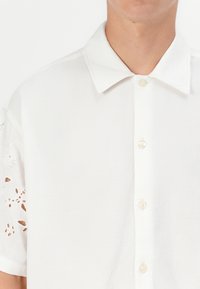 Hvid kortærmet skjorte med krave, der har intrikate udskårne broderier på ærmerne og knapdetaljer foran.