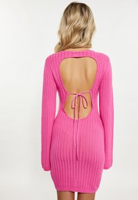 Abito a maglia rosa a coste con maniche lunghe, design con schiena scoperta e dettaglio con fiocco in vita. La texture appare morbida e aderente.