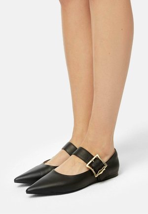 Pieds portant des chaussures plates noires à bout pointu avec de larges sangles et de grandes boucles carrées dorées sur un fond blanc uni.