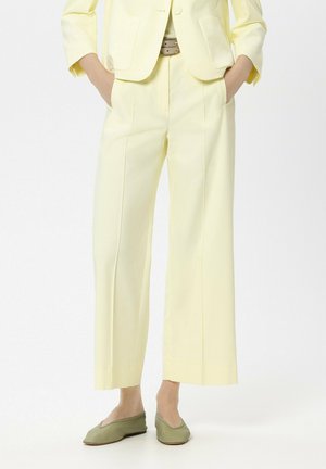 Person mit hellgelber, weit geschnittener Hose, Hände in den Taschen und olivgrünen Ballerinas vor unifarbenem Hintergrund.
