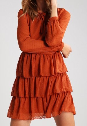 Frau trägt ein rostorangefarbenes, gestuftes Kleid mit langen Ärmeln und einer Schleife am Ausschnitt, steht mit verschränkten Armen und einer Hand nahe am Gesicht.