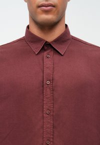 Bărbat îmbrăcat cu o cămașă burgundy, cu nasturi și guler, care își arată gâtul și partea inferioară a feței, pe un fundal simplu.