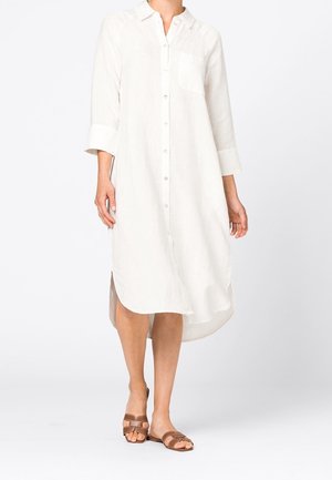 Kvinde iført en hvid, langærmet, knap-på-med-midilængde skjortekjole med brystlomme og brune åbne flade sandaler.
