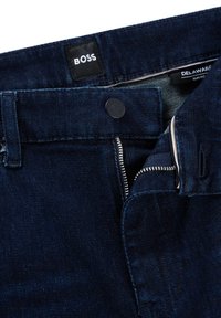 Tamsiai mėlynos džinsinės kelnės su lieknu kirpimu. Matoma prekės ženklo etiketė, metalinė saga ir užtrauktuko detalės priekyje. Minkšta tekstūra, šiek tiek išblukusi.