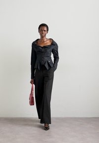 Camisa preta de botão com gola oversized, cintura peplum e cinco botões; combinada com calças pretas e uma mala rosa.