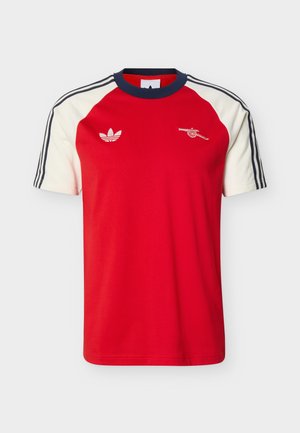 Κόκκινο και λευκό αθλητικό πουκάμισο με κοντά μανίκια, ναυτικό γιακά και ρίγες στα μανίκια, λογότυπο Adidas στο αριστερό στήθος και έμβλημα κανόνι στο δεξί στήθος.