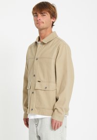 Chaqueta de lona beige con cierre de botones en la parte delantera, dos grandes bolsillos frontales y un cuello clásico. Presenta una tela texturizada y un sutil acento de logo.