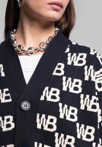 Námořnický svetr s opakujícím se bílým vzorem "WB", ribbovaný textura, V-výstřih a zapínání na dva knoflíky. Chunky stříbrný řetízkový náhrdelník.