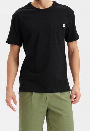 Mężczyzna w czarnej, krótkim rękawem koszulce z kieszenią na piersi oraz oliwkowo-zielonych krótkich spodenkach, stojący na białym tle.