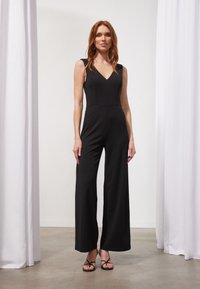 Sort jumpsuit med V-udskæring, ærmeløs design, brede ben og glat texture, suppleret med slanke sandaler. Minimalistisk stil.