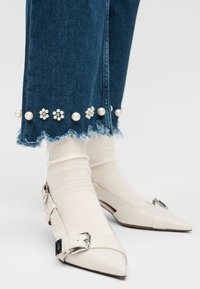 Jeans de mezclilla con bajos deshilachados y adornos de perlas, usados sobre calcetines blancos acanalados con zapatos blancos de punta puntiaguda y hebilla.