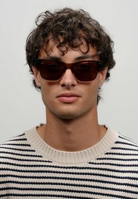 Occhiali da sole rettangolari marroni con montatura spessa; il modello indossa un maglione a righe crema e blu navy. Lo sfondo è neutro. I capelli sono ricci.