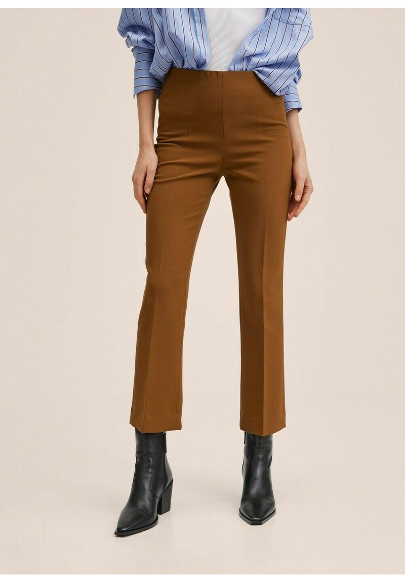 Pantalon marron ajusté avec une coupe droite, taille haute, texture lisse et détail de pli subtil. Porté avec des bottines noires et un haut à rayures bleues.