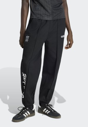 Person trägt schwarze Adidas-Sporthose mit Liverpool-FC-Wappen und weißem Schriftzug am linken Bein, kombiniert mit schwarzen Adidas-Sneakern mit weißen Streifen.
