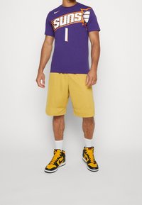 Lilla Nike Suns-trøje med hvidt nummer 1, gule shorts og sorte og gule højtop-sneakers. Hvide ankelsokker fuldender outfittet.