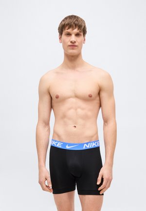 Jeune homme debout devant un fond uni, portant un boxer noir Nike avec une ceinture bleue et le logo Nike.