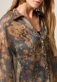 Camicia a stampa floreale in tessuto trasparente, con tonalità di blu e marrone, colletto decorato, chiusura con bottoni e maniche lunghe.