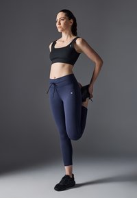 Crni atletski crop top s širokim naramenicama; mornarsko plave tajice visokog struka s elastičnom trakom za podešavanje. Model izvodi stojeće istezanje.